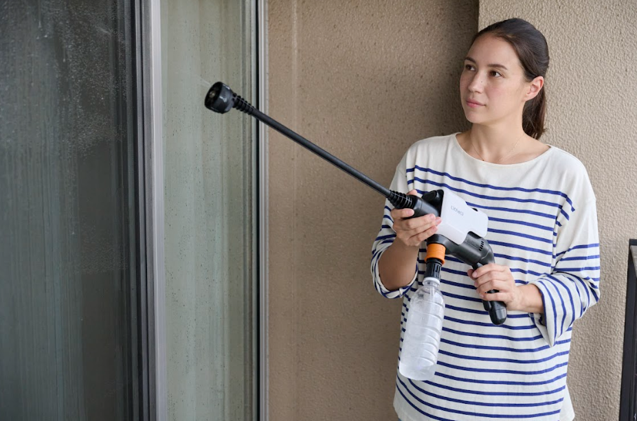 バルコニーでポータブル高圧洗浄機を両手で構え窓を洗浄するボーダーシャツの女性
