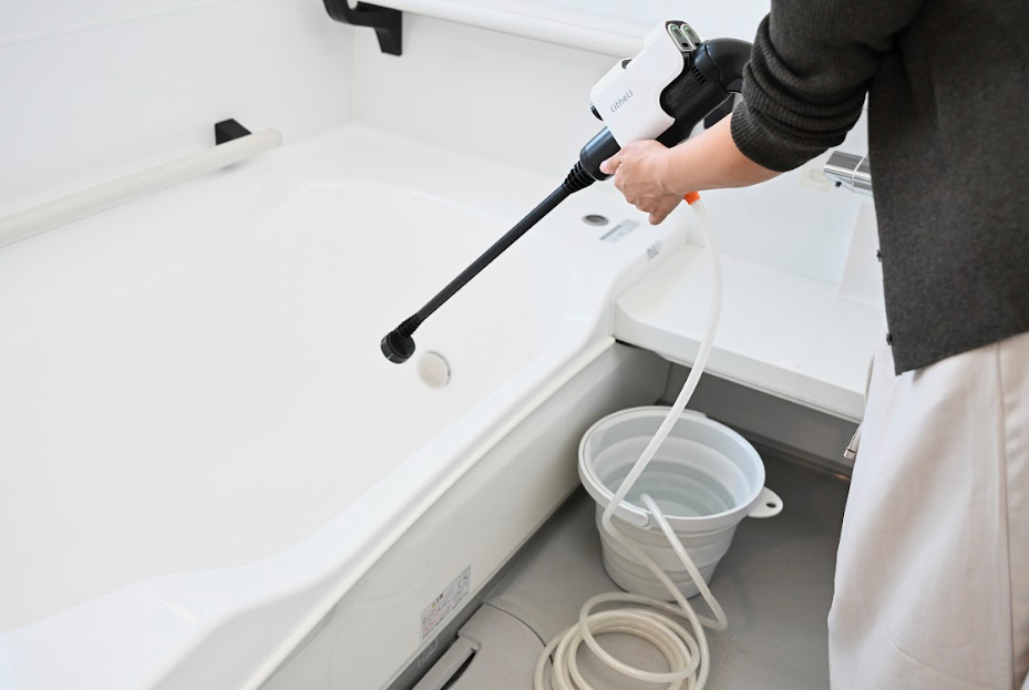 バケツの水を吸い上げるホースをセットしたポータブル高圧洗浄機で浴槽を洗う手元アップ