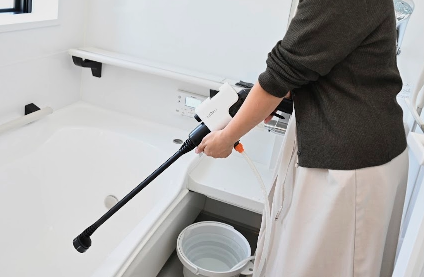 バスルームでバケツの水をポータブル高圧洗浄機で浴槽に向けて使用する女性の手元